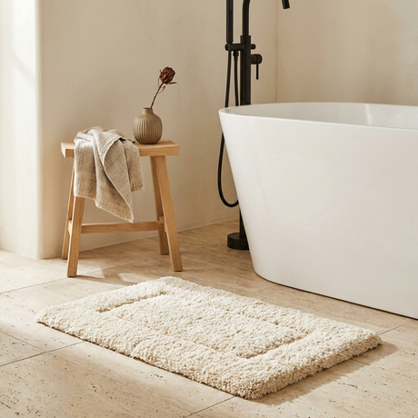 Cream tufted bath mat on travertine tile beside a freestanding tub, oak stool with folded linen towel