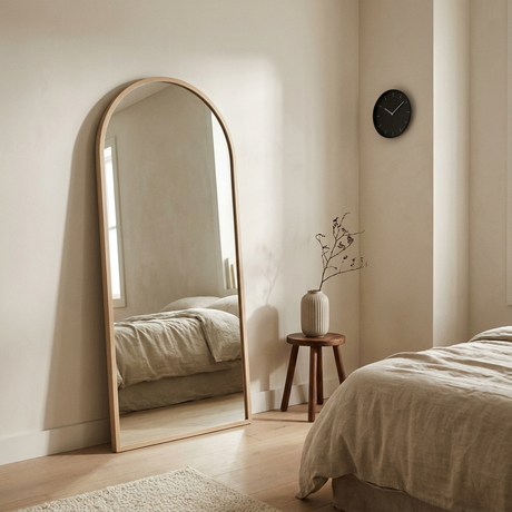 Full-length arched oak-framed mirror leaning against a warm off-white plaster wall in a bedroom corner, matte-black wall clock nearby